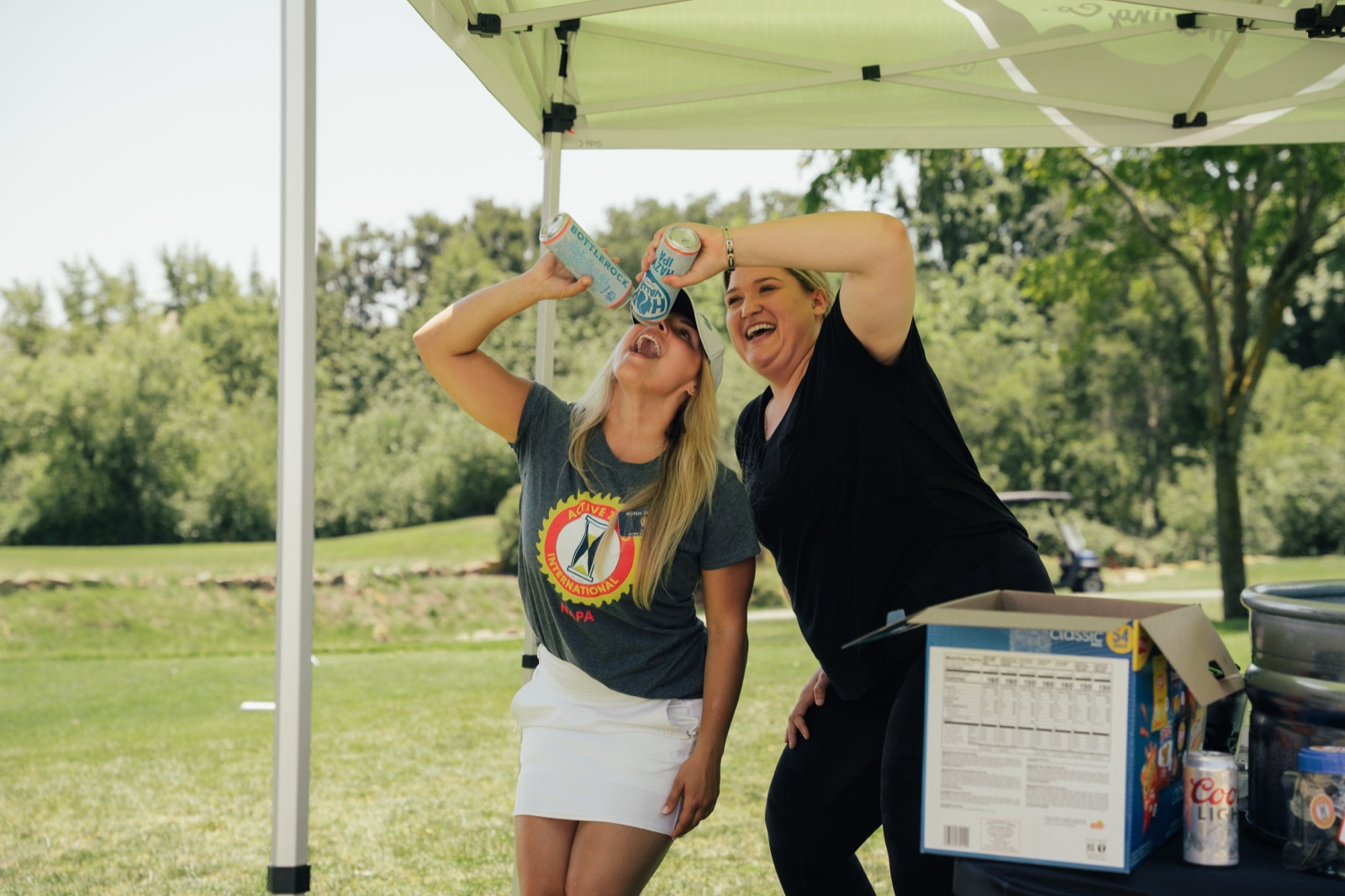 Two smiling women under a green canopy adjusting cornhole boards, one in a logo shirt and the other in black.