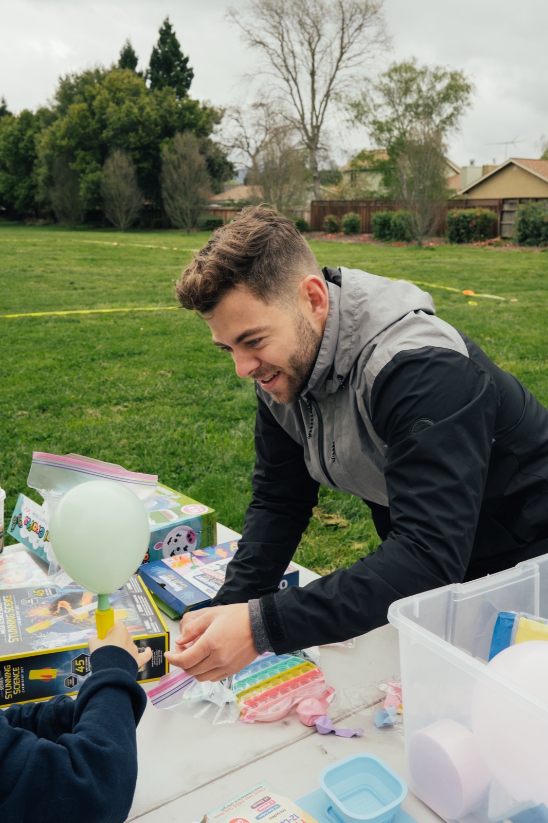 A young adult volunteer smiling while assisting a child with an Easter craft at an outdoor table with supplies.