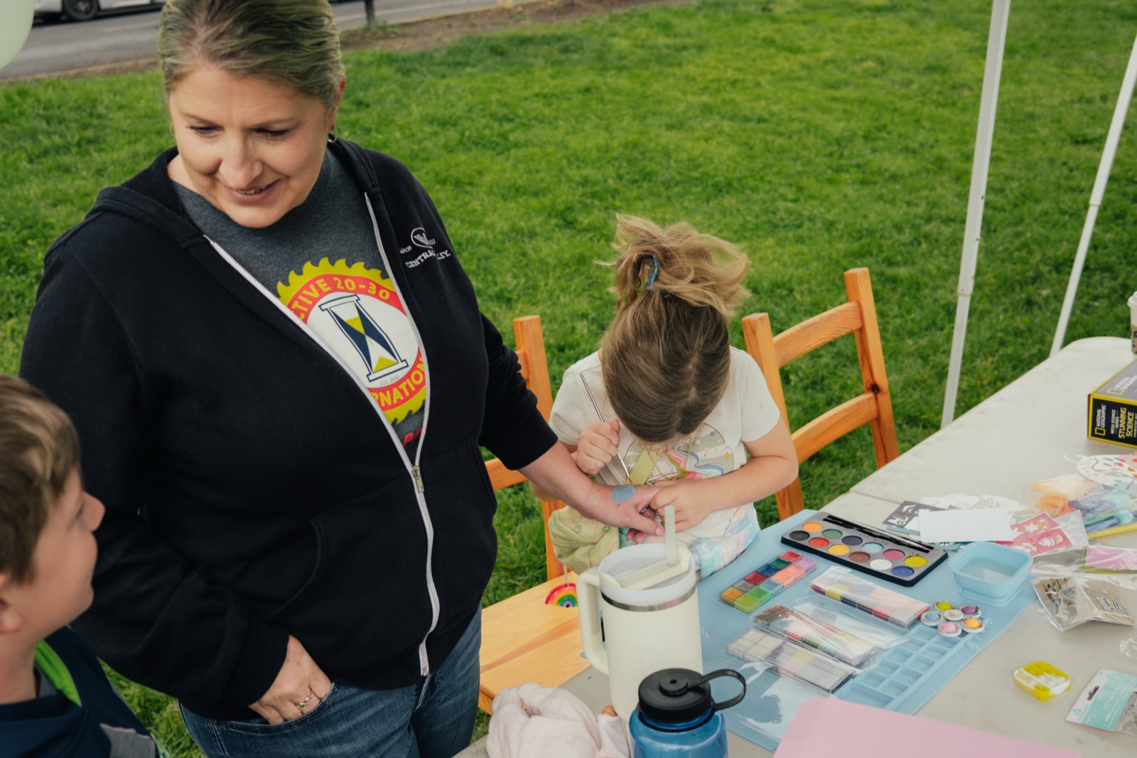 A smiling volunteer in a Napa Active club jacket assisting a child with an Easter craft activity.