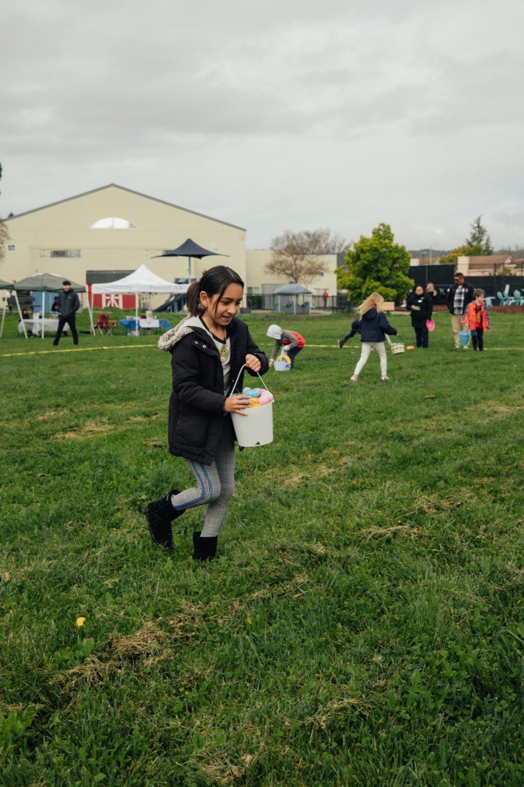 A child joyfully running across the grassy field with a filled Easter basket, with volunteers and other participants visible in the background.