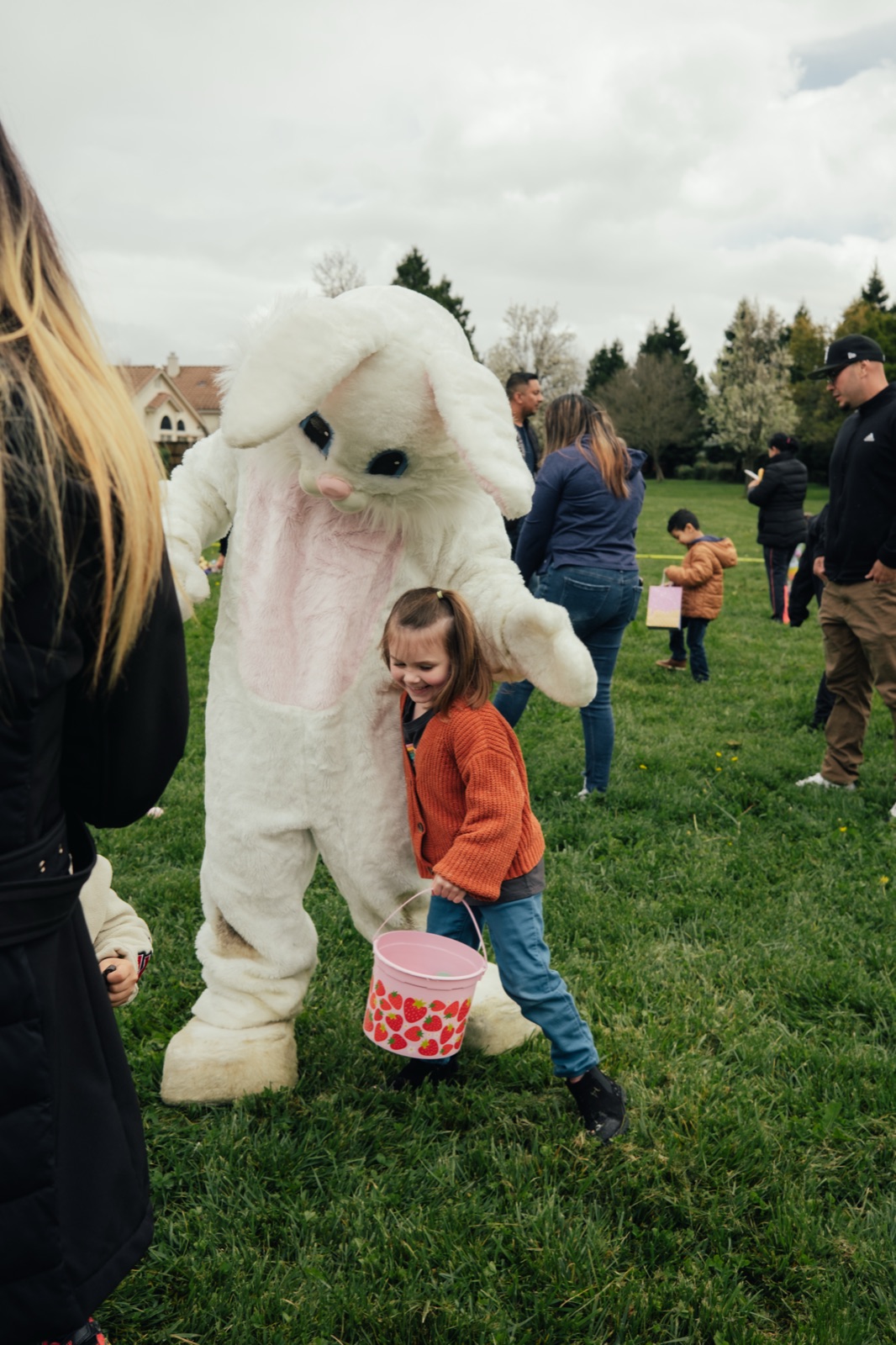 Small child holding pink Easter basket greeting Easter bunny mascot on grass.